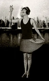 A black-and-white 1920s photograph of a woman in a bathing suit and swim cap posing beside a lake, one arm raised